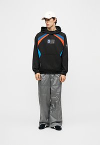 Jeune homme portant un sweat à capuche noir avec des accents bleu et orange, une casquette blanche, un pantalon large gris et des chaussures noires, debout devant un fond uni.