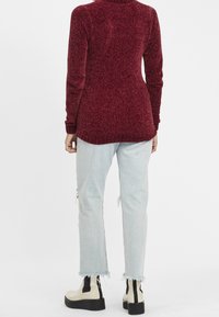 Bordeaux gebreide trui met hoge hals en geribbelde manchetten, in combinatie met lichtblauwe distressed jeans en crèmekleurige enkellaarsjes met zwarte accenten.