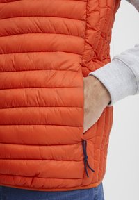 Puffet orange vest med vandrette kviltede sektioner, sidelommer og snørelukning. Indersiden synlig gennem ærmet.