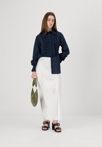 Cămașă bleumarin cu volane, cu nasturi în față, asociată cu o fustă maxi din satin alb. Modelul ține o poșetă verde și poartă pantofi cu toc negri.