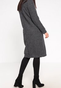 Cappotto lungo a righe bianche e nere con taglio dritto, maniche lunghe, spacchi laterali e una finitura testurizzata, abbinato a leggings neri aderenti e stivaletti alla caviglia.