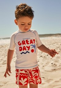 Litet barn klädd i en vit rash guard med blå detaljer och röd grafisk text, tillsammans med röda badshorts med ett kraftigt vitt mönster, på stranden.