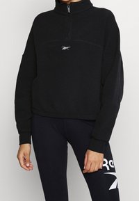 Sweat-shirt zippé en polaire noire avec un col montant, présentant une couture frontale et un logo blanc sur la poitrine. Texture douce et coupe décontractée.