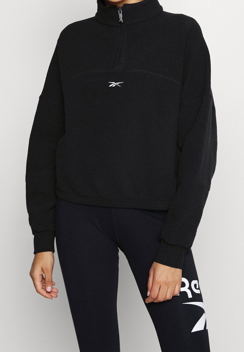 Sweat-shirt zippé en polaire noire avec un col montant, présentant une couture frontale et un logo blanc sur la poitrine. Texture douce et coupe décontractée.