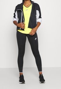 Giacca Nike bianca e nera su un top giallo brillante, abbinata a leggings neri con un logo. Scarpe da atletica nere completano l'outfit.