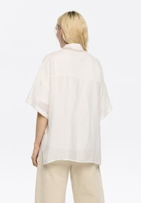 Camicia bianca leggera e oversize con tessuto strutturato, maniche corte e spacco posteriore. Presenta sottili motivi a griglia e spacchi laterali.