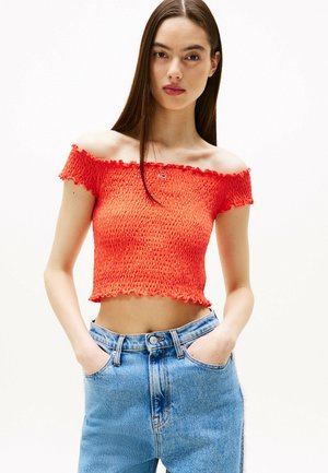 Top naranja recortado, con escote Bardot y textura fruncida, con bordes en volantes; combinado con jeans de tiro alto en azul claro.