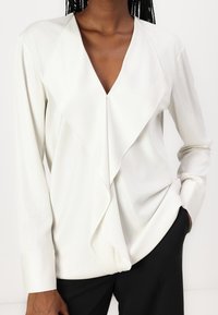 Camicia bianca con scollatura a V profonda e frontale drappeggiato. Maniche lunghe e texture liscia, abbinata a pantaloni neri.