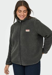 Szara kurtka polarowa z wysokim kołnierzem i zamkiem błyskawicznym z przodu. Na lewej piersi znajduje się mała prostokątna naszywka z logo marki. Miękka, teksturowana powierzchnia.