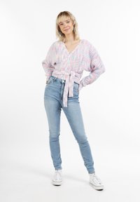 Pastel krāsas apģērbs ar plašām piedurknēm, sasieta jostasvietā. Apvienots ar gaiši zilām šaurām biksēm un baltām sporta apaviem. Vienkārša fona.