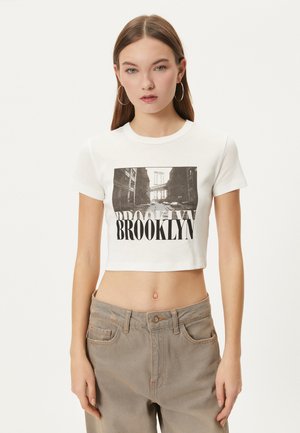 Camiseta corta blanca con un estampado gráfico negro de una escena de Brooklyn y el texto en negrita "BROOKLYN" debajo. Tejido de algodón suave.
