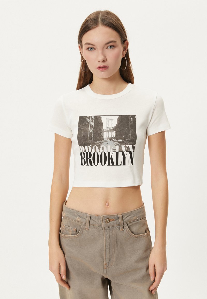 Hvid cropped t-shirt med et sort grafisk print af en Brooklyn-scene med fed tekst "BROOKLYN" nedenunder. Blødt bomuldsstof.