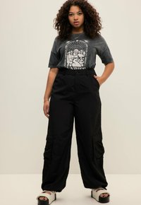 Zwarte cargo broek met zakken, gecombineerd met een anthracietgrijs T-shirt met grafische print in wit. Het model draagt witte slippers.