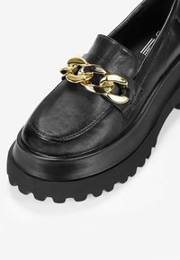 Mocassino in pelle nera con suola piatta spessa, caratterizzato da un accento di catena dorata sulla tomaia e suola in gomma testurizzata.