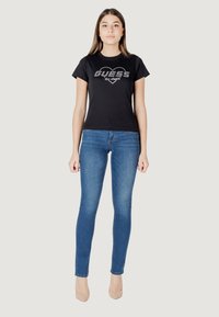 Zwarte katoenen t-shirt met zilveren "GUESS Los Angeles" tekst. Gecombineerd met blauwe skinny jeans en beige hakken, staat het model recht van voren.