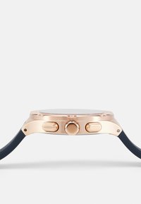 Boîtier de montre en métal doré avec deux boutons-poussoirs et une couronne, fixé à un bracelet en silicone bleu foncé. Cadran en verre rond visible.