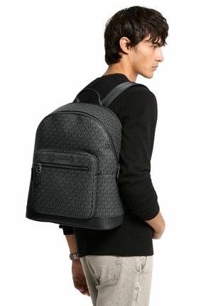 Giovane uomo con maglione nero e jeans chiari, che porta uno zaino nero con fantasia su una spalla, di profilo su sfondo bianco.