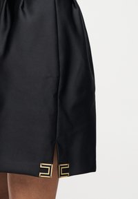 Μαύρη σατέν φούστα με μικρή σχισμή μπροστά, διακοσμημένη με δύο χρυσά γεωμετρικά μεταλλικά στοιχεία κοντά στο τελείωμα, φορεμένη από άτομο με σκούρο δέρμα.