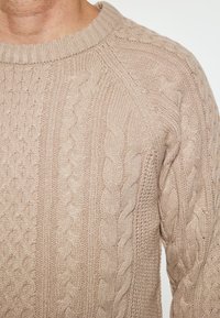 Beige gebreide trui met gestructureerde kabelpatronen, ribbelhalslijn en een zachte afwerking. Close-up weergave benadrukt de ingewikkelde details.