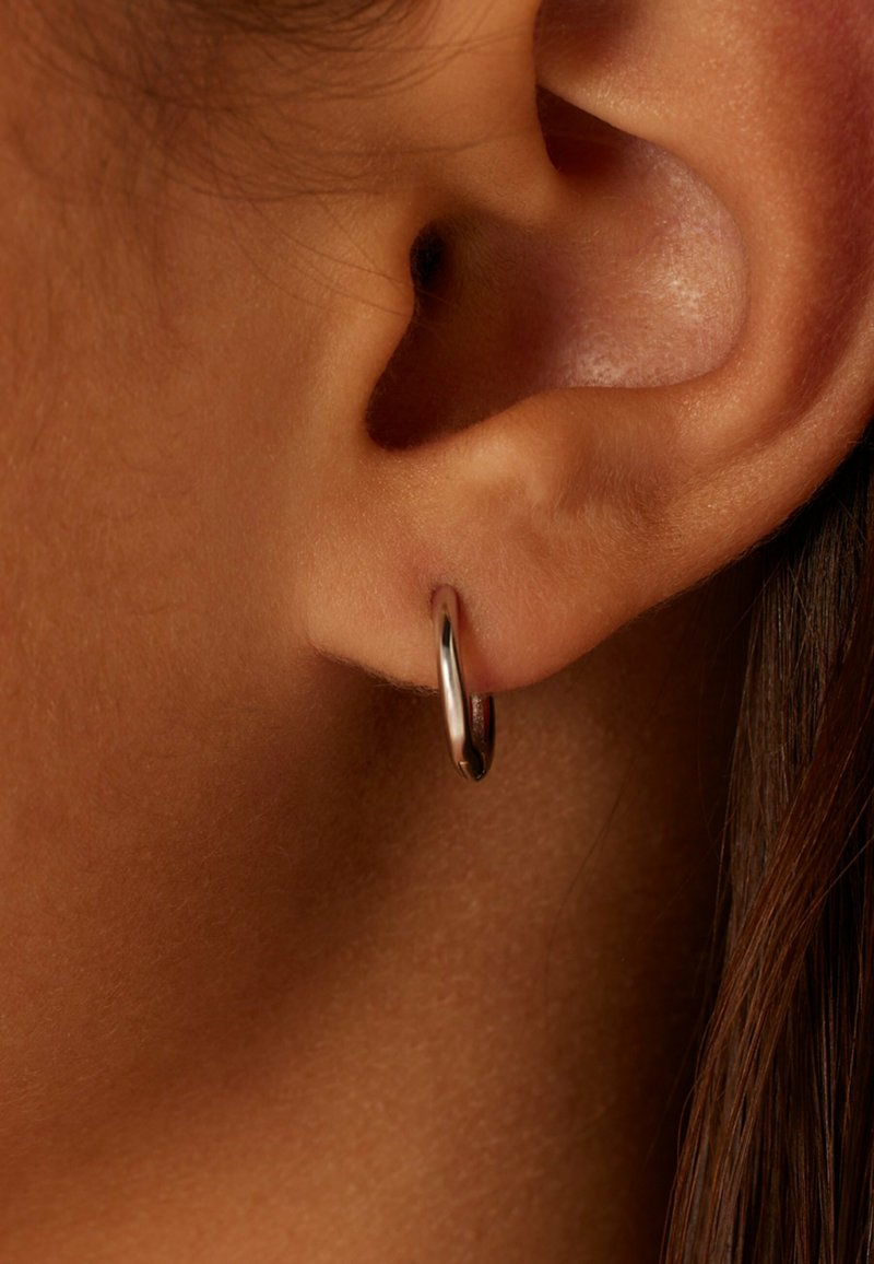 Kleine silberne Creolen, die am Ohrläppchen präsentiert werden, mit einer glatten, spiegelnden Oberfläche und einem kreisförmigen Design ohne zusätzliche Verzierungen.