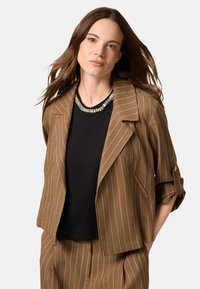 Braun gestreifter Blazer mit lässiger Passform, darunter ein schwarzes Oberteil und ein dekoratives Perlendetail am Halsausschnitt, glatte Stoffstruktur.