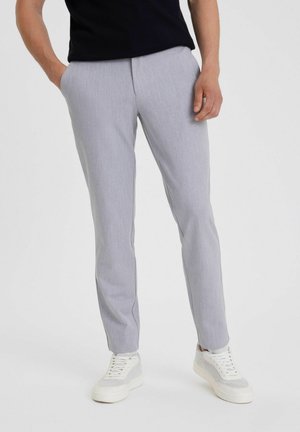 Man met lichtgrijze slim-fit broek en witte sneakers, staand met één hand in de zak tegen een effen achtergrond.