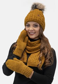 Gebreide mosterdkleurige muts met een bont pompon, bijpassende sjaal en wanten. Gedetailleerd patroon, geribbelde manchetten op de wanten en de muts. Warm, gezellig winters set.