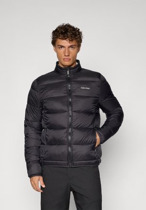 Svart dunjakke med høy krage, glidelås foran og horisontal quiltet design. Har Calvin Klein-logo på brystet og sidelommer.