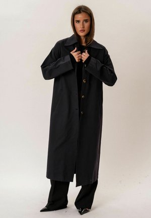 Młoda kobieta w długim czarnym płaszczu z guzikami, stojąca na tle jednolitego jasnego tła, poprawiająca kołnierz.