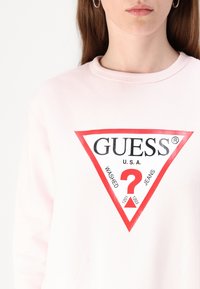 Sweatshirt rose clair présentant un grand logo triangulaire rouge avec les mots "GUESS", "WASHED" et "JEANS", et un point d'interrogation rouge au centre.