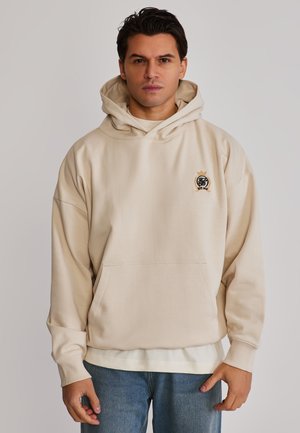 Homme portant un sweat à capuche beige avec poche avant et emblème noir, assorti à un jean bleu clair, debout devant un fond uni.