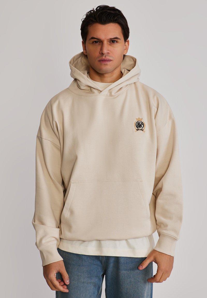 Homme portant un sweat à capuche beige avec poche avant et emblème noir, assorti à un jean bleu clair, debout devant un fond uni.