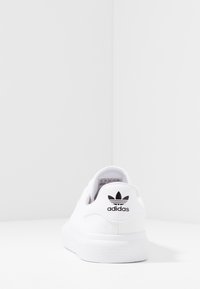 Biała tenisówka na płótnie z teksturowaną gumową podeszwą. Czarne logo Adidas na pięcie i zaokrąglony kształt noska. Minimalistyczny styl i wykonanie.