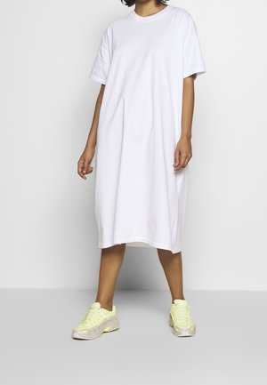 Osoba ubrana w luźną białą sukienkę T-shirtową do kolan i neonowożółte sneakersy, stojąca na tle jednolitego białego tła.