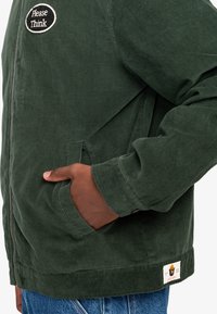 Grøn kordfløjljakke med et rundt sort og hvidt patch med teksten "Please Think", med sidelommer og lynlås-lukning.