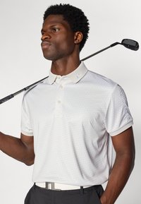 Polo shirt blanc avec un motif géométrique subtil, col gris et logo sur l'épaule, associé à un pantalon noir et une ceinture blanche. Club de golf sur l'épaule.