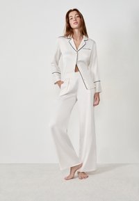 Femme debout pieds nus, portant un ensemble de pyjama en satin blanc avec des bordures noires, une main dans la poche, devant un fond clair uni.