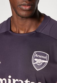 Maillot de sport violet foncé, avec un col rond, des épaules texturées et un écusson d'Arsenal FC en blanc avec un graphique de canon.