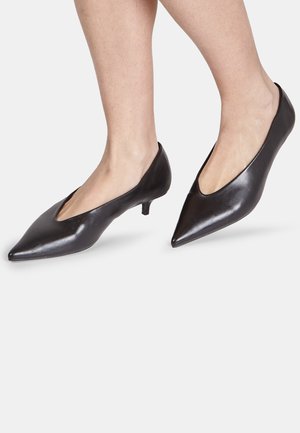 Scarpe nere in pelle con tacco kitten a punta indossate a piedi nudi su uno sfondo chiaro uniforme.