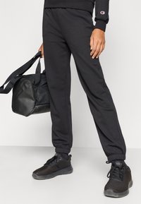 Μαύρο παντελόνι φόρμας με ελαστική μέση και εφαρμοστά τελειώματα, κατασκευασμένο από μαλακό ύφασμα. Το άτομο κρατά μια μαύρη τσάντα duffle με το ένα χέρι, φορώντας μαύρα αθλητικά παπούτσια.