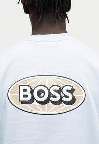 Personne à la peau foncée et aux dreadlocks portant une chemise blanche avec un logo ovale beige et noir indiquant "BOSS" dans le dos.
