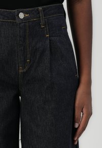 Dunkelblaue Jeanshose mit hoher Taille, mit Falten, kontrastierenden gelben Nähten und einem Metallknopfdetail am Bund.