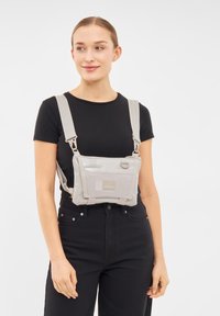 Borsa a tracolla grigia chiara realizzata in tessuto resistente, con spallacci regolabili, tasca frontale e logo del marchio, indossata sopra una maglietta nera.