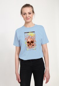 Svetle modré tričko s krátkým rukávem, na němž je grafický design lebky s oranžovými a žlutými akcenty, umístěný na stylizovaném pozadí.