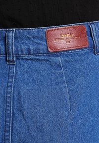 Pantalon en denim bleu avec une étiquette en cuir marron embossée de texte. Le tissu présente une texture lisse et un design à coupe classique.