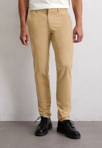 Beżowe chinosy o slim fit, z płaskim przodem, bocznymi kieszeniami i gładką strukturą, zestawione z czarnymi butami na sznurówki.