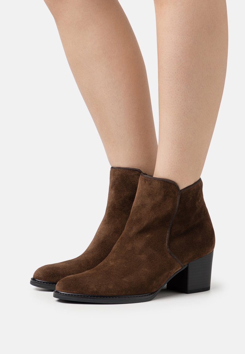 Bottes marron en daim à talons carrés portées sur des jambes nues sur fond blanc uni.