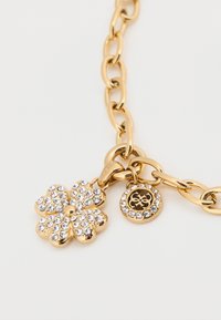 Βραχιόλι από χρυσή αλυσίδα με μοτίφ λουλουδιού διακοσμημένο με διαφανείς κρυστάλλους και κυκλικό μενταγιόν με λογότυπο, και τα δύο σε λεία αλυσίδα.