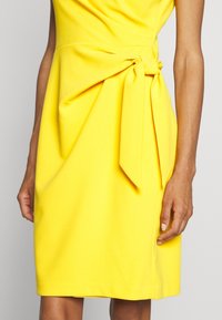 Gul klänning med draperad halsringning och sidoknytning. Gjord av ett mjukt tyg, har den en Figursydd silhuett och knälång fåll.