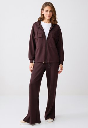 Jeune femme portant un sweat à capuche marron foncé zippé avec poches avant, pantalon large assorti, T-shirt blanc et baskets beiges, debout de manière détendue.